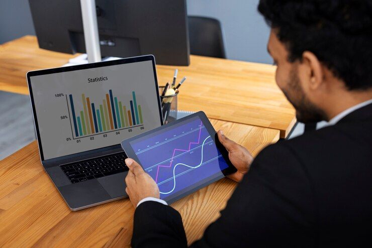 Business professional analyzing currency market statistics using a tablet and laptop showing bar and line charts.