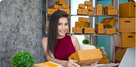 Female small business owner holding a package while working on a laptop, with shelves full of parcels in the background.