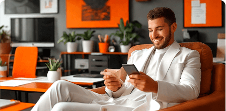 Smiling man in white suit sitting in a modern orange-accented office, using his phone and holding a credit card.